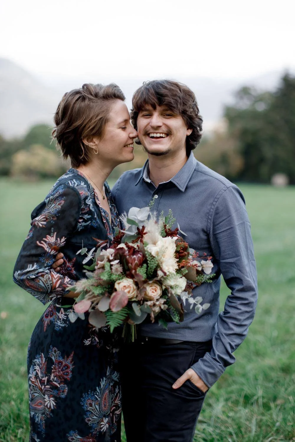 A happy couple posing outdoors on a cloudy day, with the woman holding a bouquet of flowers. The woman is leaning in to kiss the man on the cheek as he smiles.