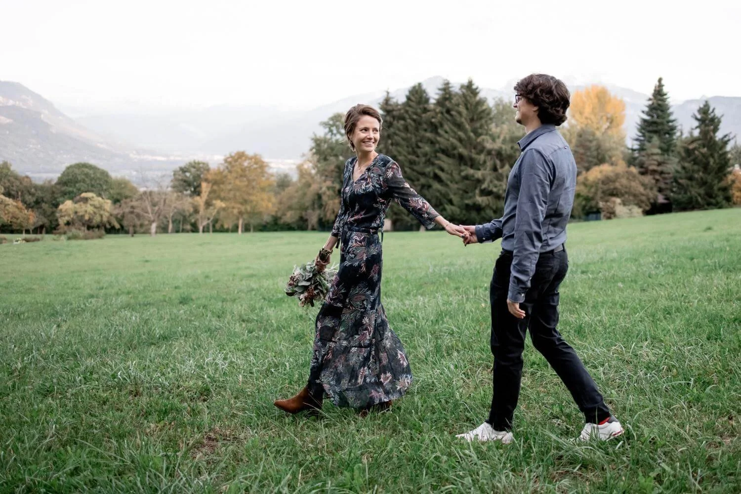 A woman in a long floral dress holding a bouquet and a man in a blue shirt and black pants holding hands in a grassy field with trees and mountains in the background.