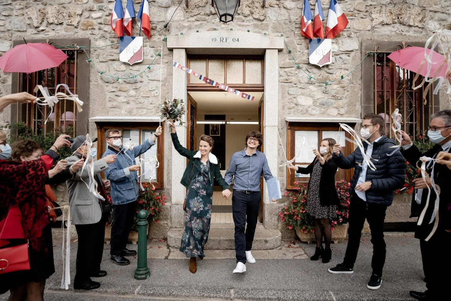 A couple leaves a building holding hands as they are cheered by friends and family during a celebration outside a stone building decorated with French flags and ribbons. The people are wearing masks.