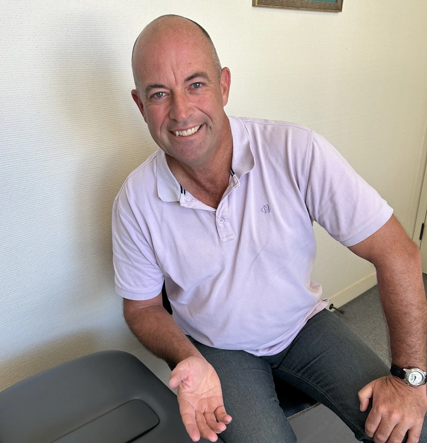 A smiling man with a shaved head, wearing a light purple polo shirt, sitting in a clinical setting. He is assisting a patient.