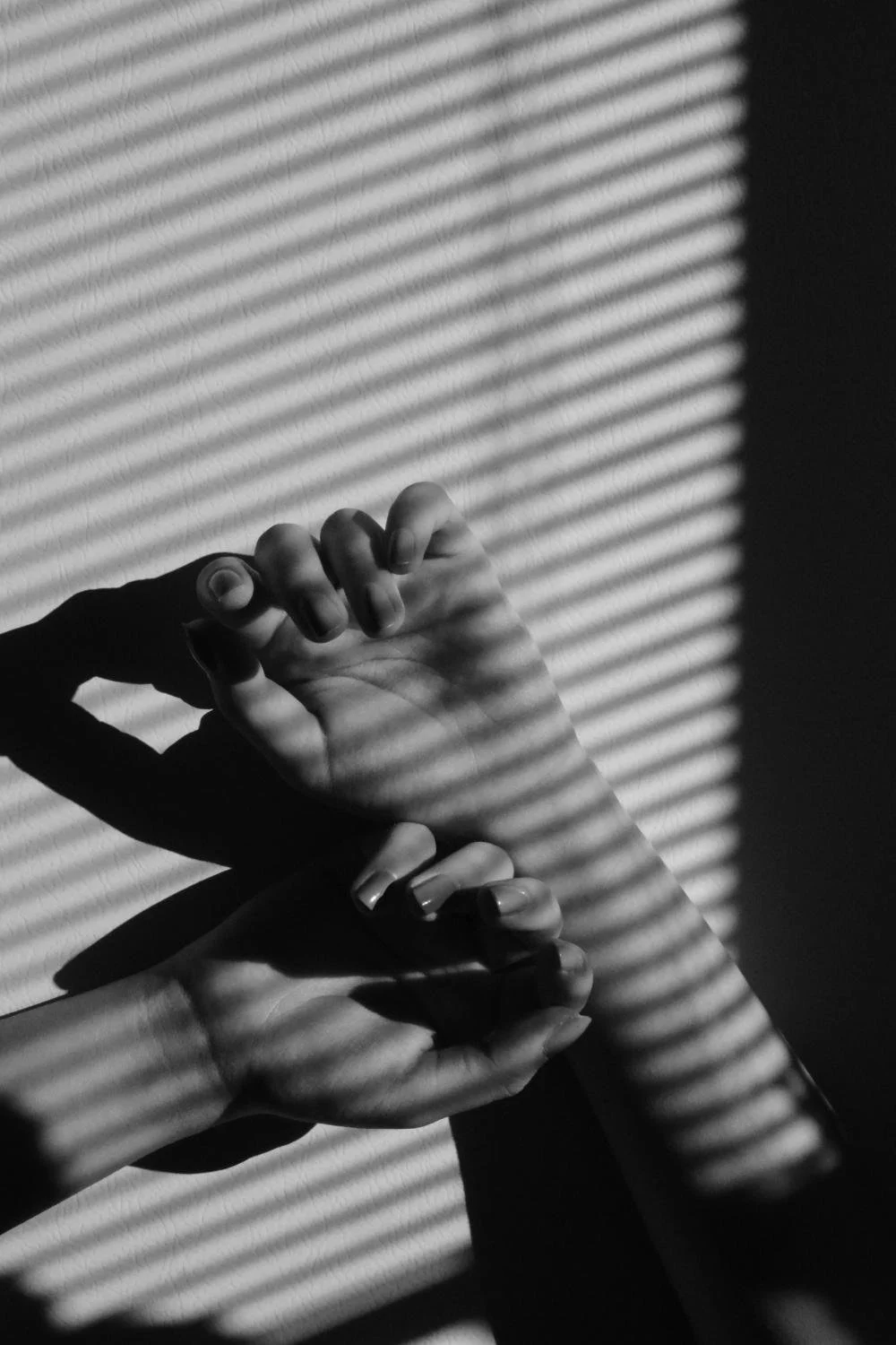 Black and white photo of hands stretching towards a wall with horizontal blinds casting striped shadows.