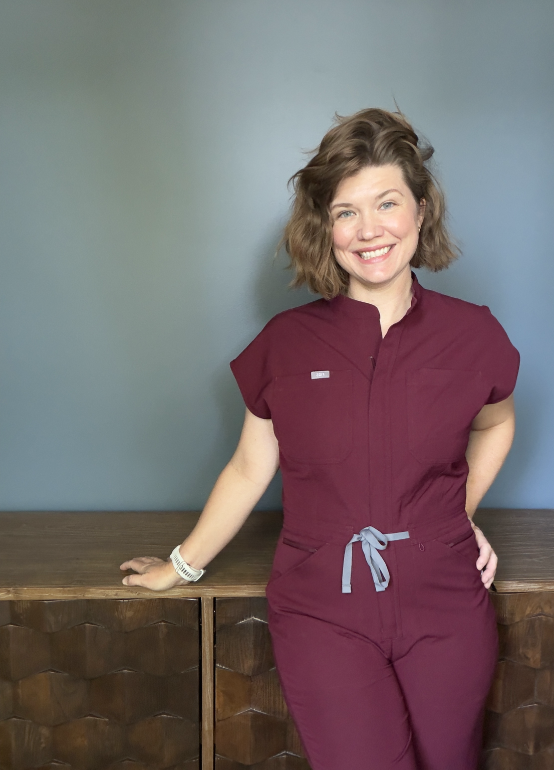 Woman with brown hair wearing maroon medical scrubs, smiling, standing in front of a teal wall, with one hand resting on a wooden cabinet.