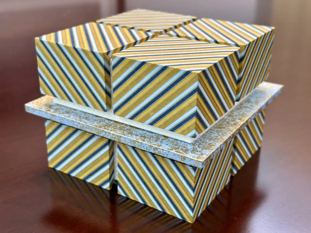 A decorative geometric sculpture with a striped pattern in yellow, blue, black, and white, placed on a wooden surface.