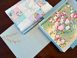 A decorative box with artificial pink and white flowers, a pair of pearl earrings, and a quilted fabric piece, all on a wooden surface.