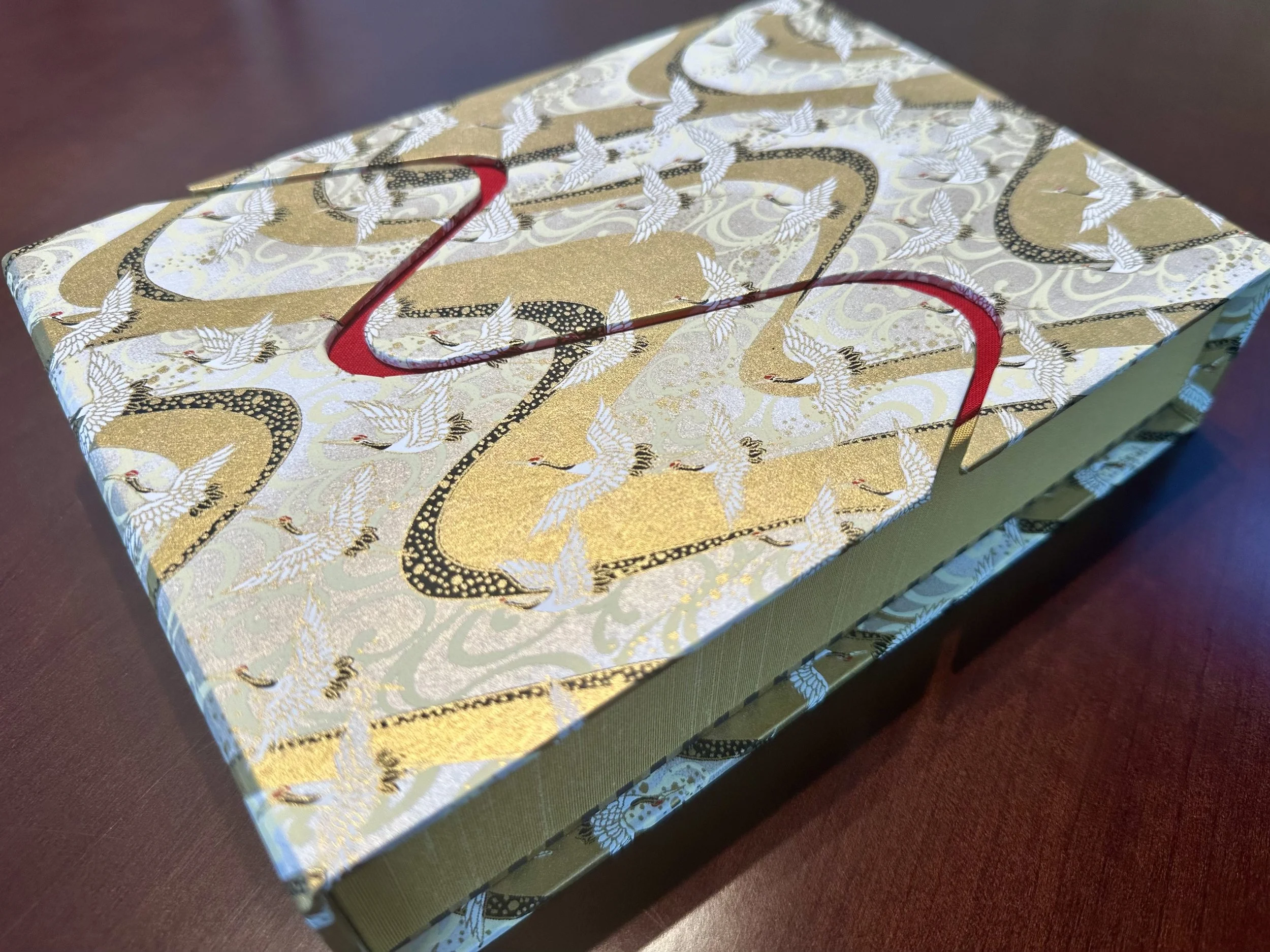Decorative gift box with a pattern of white cranes and gold accents, tied with a red ribbon, resting on a dark surface.