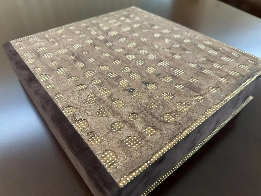 A closed box with a textured brown lid featuring a pattern of evenly spaced circular perforations with small white dots, resting on a dark surface.