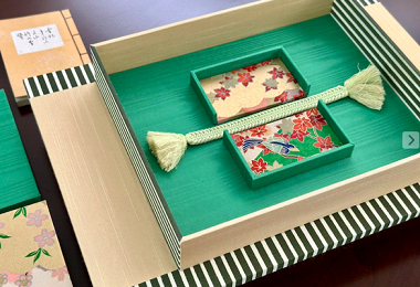 A decorative gift box contains two small boxes with holiday-themed patterns, a tassel-shaped object, and a themed paper. The box is placed on a striped surface.