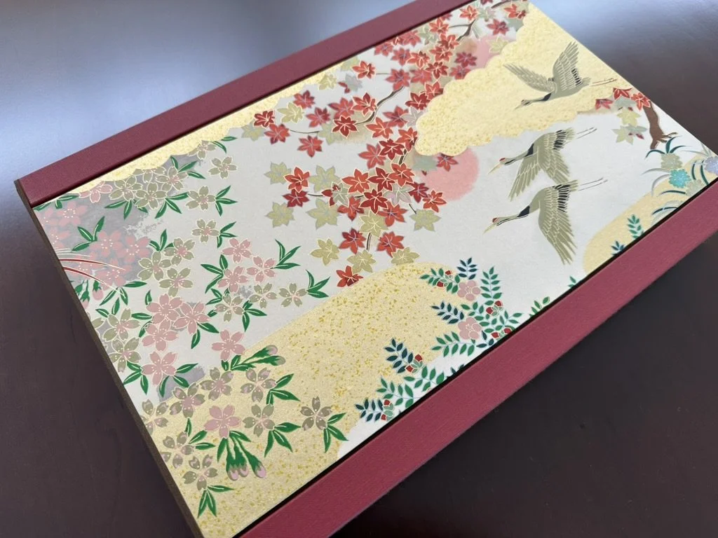 Decorative rectangular box with a traditional Japanese scene featuring cherry blossoms, cranes, and a partly cloudy sky with a red sun.