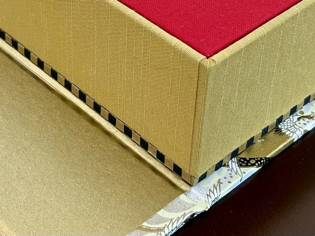 Close-up of the corner of a beige upholstered piece of furniture with a red cushion on top. The furniture has a black and yellow checkered trim and is placed on a patterned fabric with butterflies, on a tiled floor.