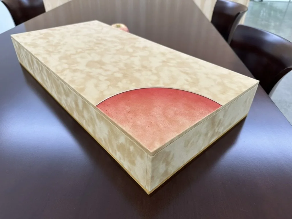 A board game box with a cream-colored marbled exterior and a small red semi-circular section in the corner, placed on a dark wooden table.