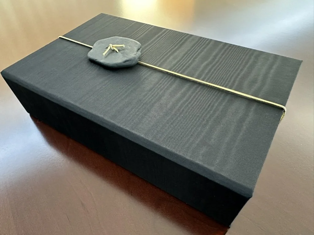 A black coffin with a gray pillow on top, decorated with a beige rope and placed on a dark wooden surface.
