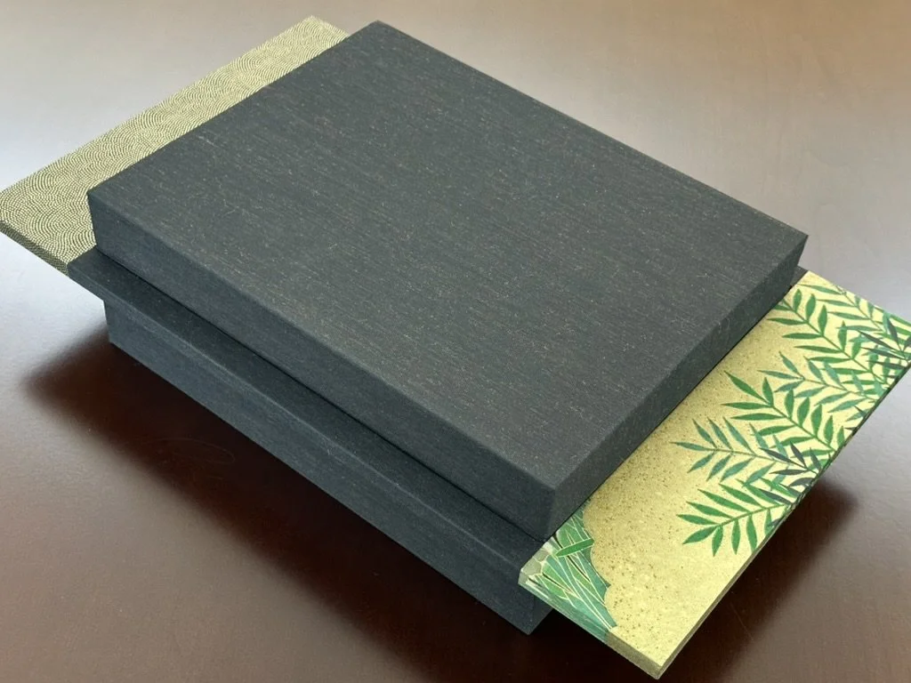 A black box with textured surface resting on decorative paper with green leaves on a gold background.