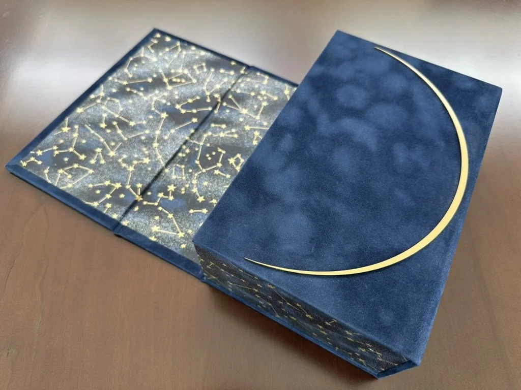 A navy blue box with a gold crescent moon design on top and a constellation pattern on the inside, resting on a wooden surface.