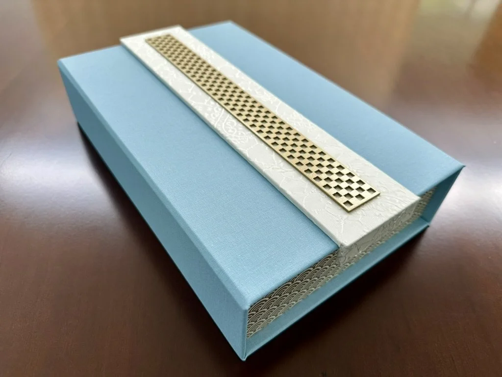 A decorative blue box with a white textured lid and a gold strip, placed on a wooden surface.