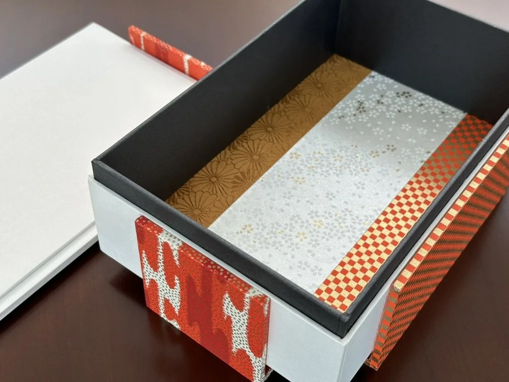 Open decorative gift box with patterned wrapping paper inside, next to a closed white box on a dark surface.