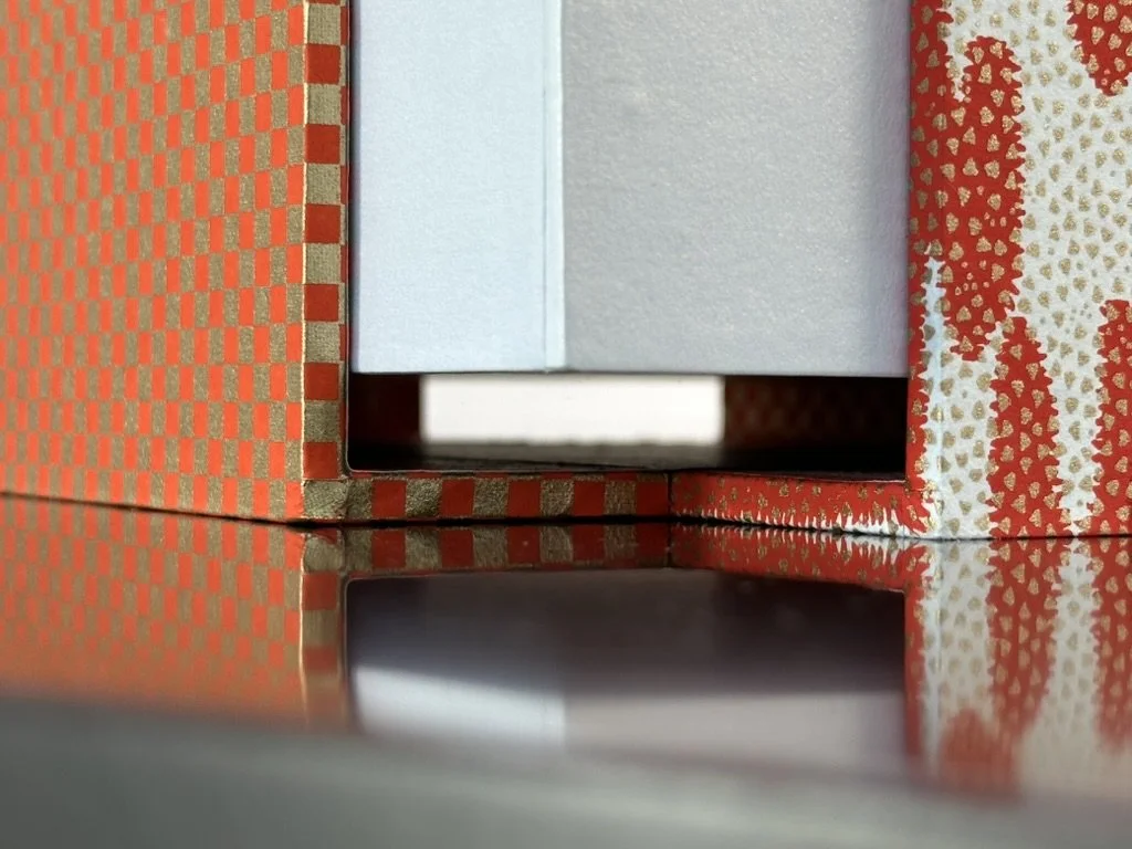 Close-up of a cardboard box with red and gold checkered pattern, showing a small gap revealing a white object inside.