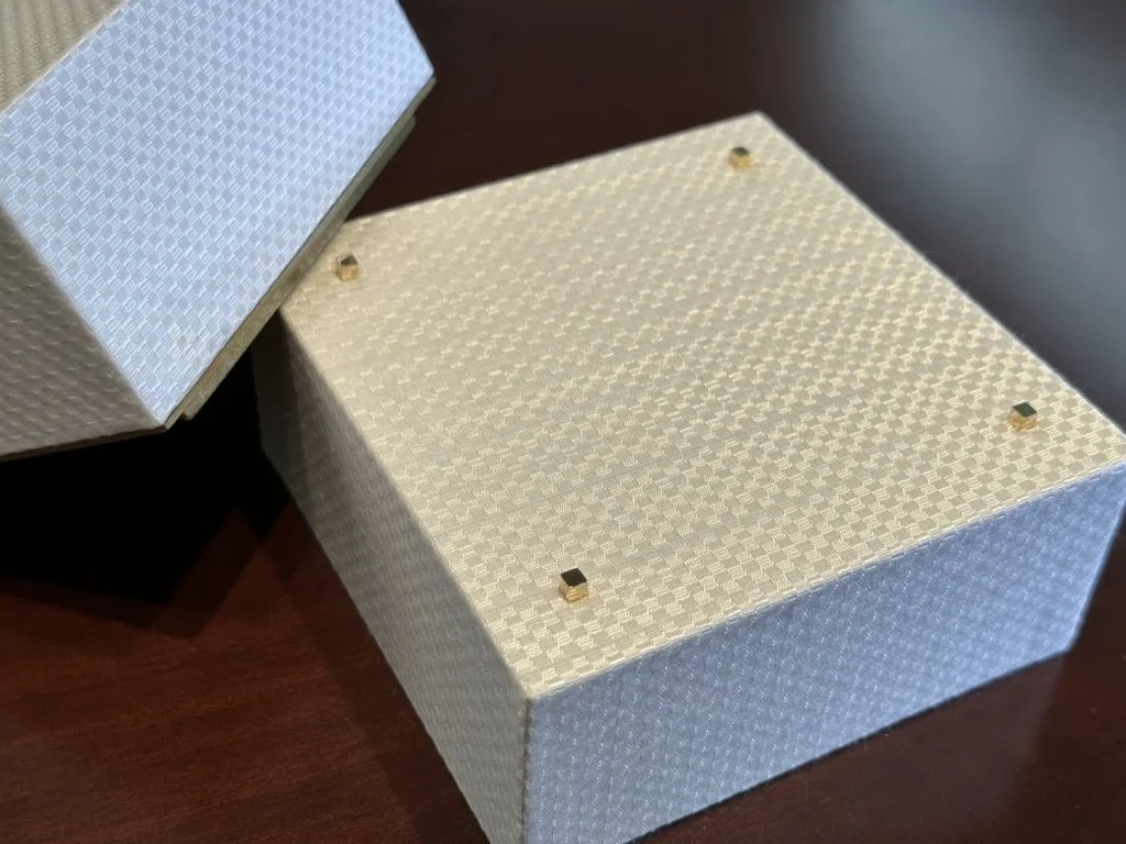 A closed, square jewelry or gift box with a textured, cream-colored surface and four small black and gold decorative accents on the top, resting on a dark wooden surface with another similar textured box partially visible nearby.