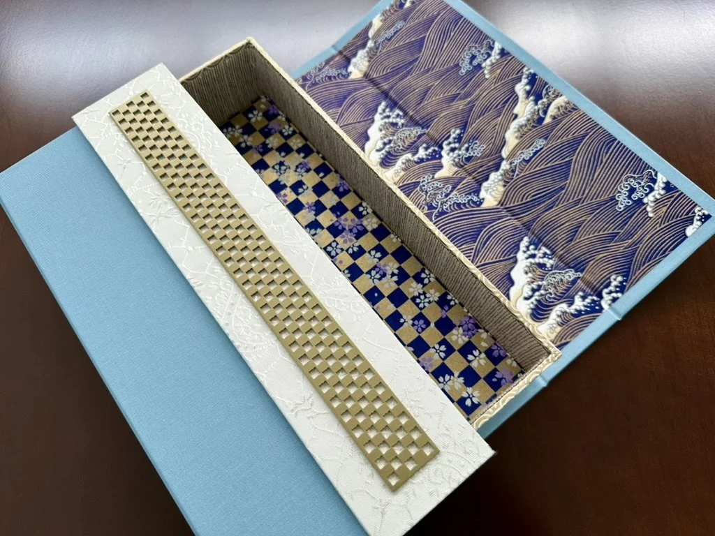 Open decorative box with patterned interior and a long metallic grid object inside.