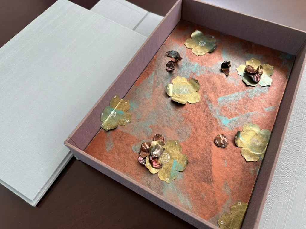 A pink and orange origami paper box partially open, containing folded and crumpled pieces of shiny metallic paper in gold, copper, and rose gold colors, arranged on a textured orange and teal paper background.