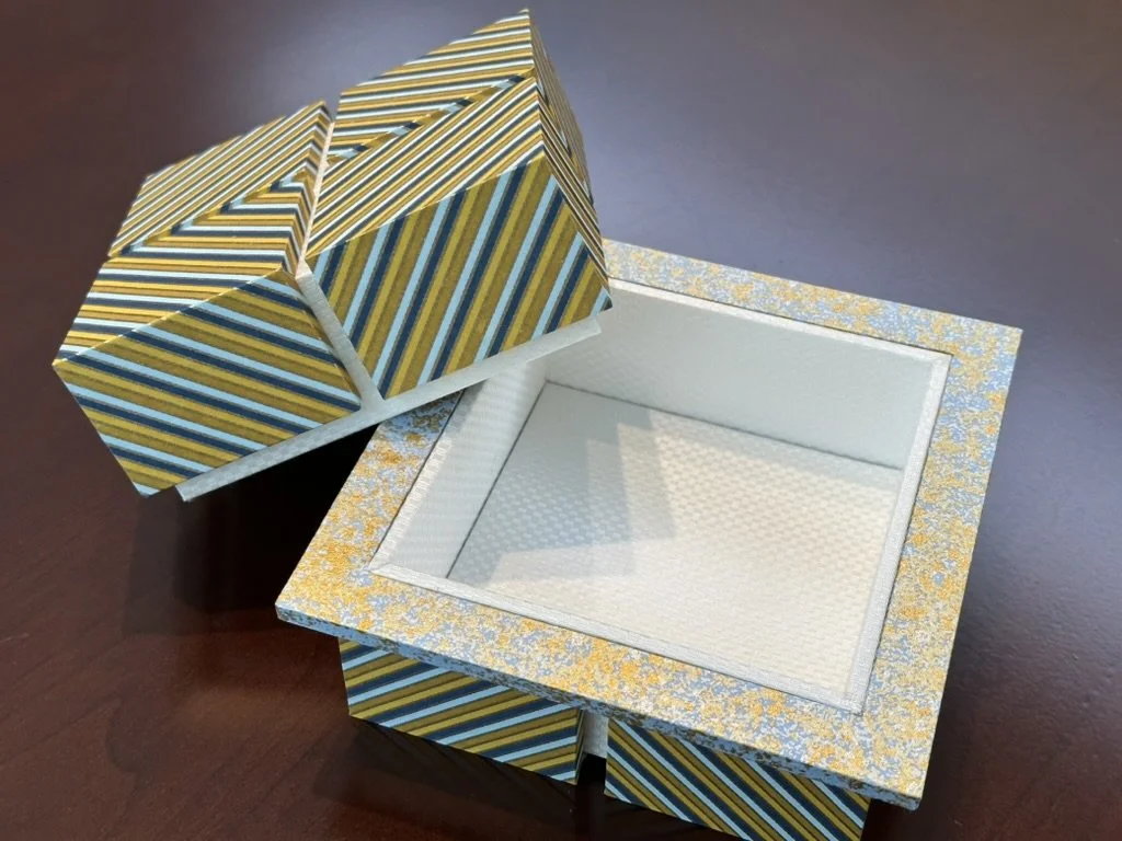 An open decorative box with a lid resting beside it. The box has a floral pattern, and the lid has a striped pattern with yellow, blue, and black stripes.