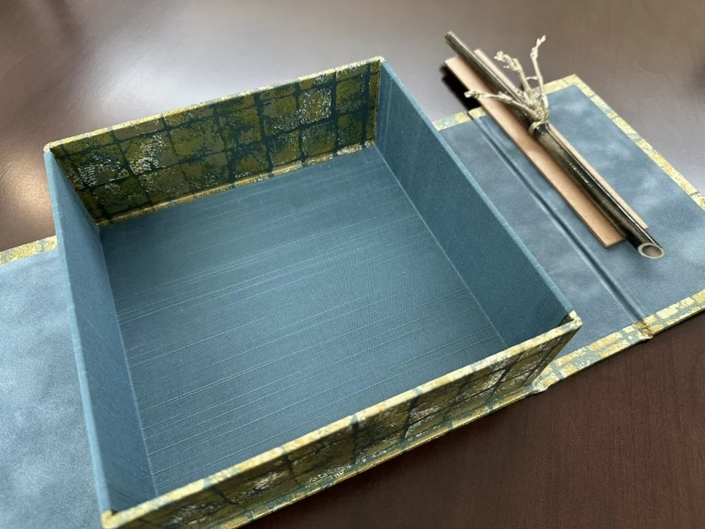 Open decorative box with blue interior on table, paired with a rolled scroll tied with string on a matching surface