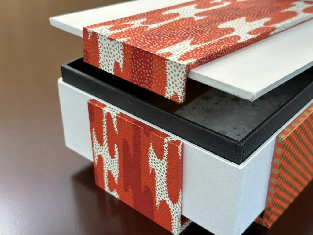 Close-up of a white box with a black interior, decorated with red and white patterned wrapping paper on the lid and front, sitting on a dark wooden surface.