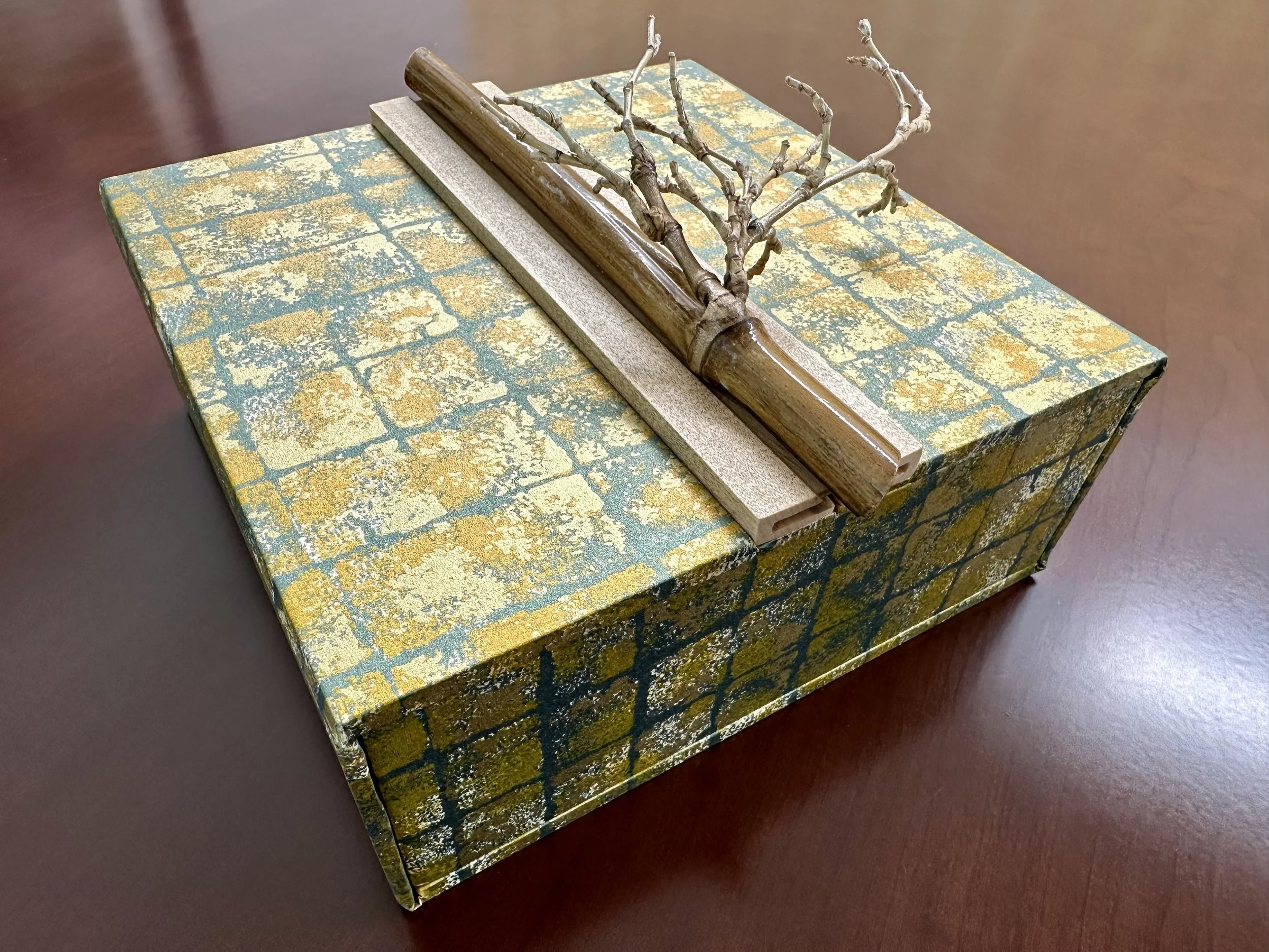 A decorative box with a square pattern in yellow, blue, and gold colors, featuring a small, dried, leafless branch and a wooden tool on top.