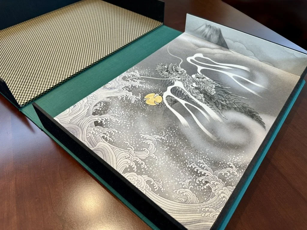 Open presentation box with traditional Japanese artwork of waves, flowers, and a mountain, along with textured fabric swatches inside.