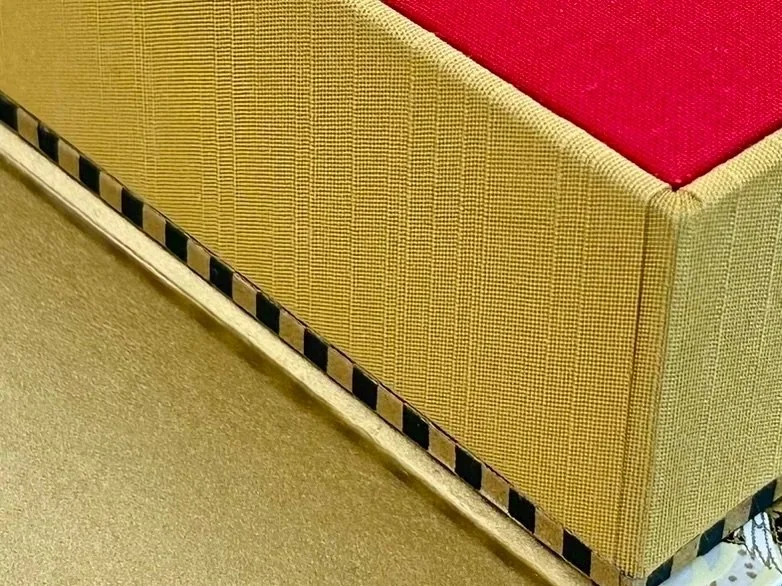 Close-up of a yellow fabric sofa with a red cushion on top and black-and-gold decorative trim along the bottom edge, next to a beige carpeted floor.