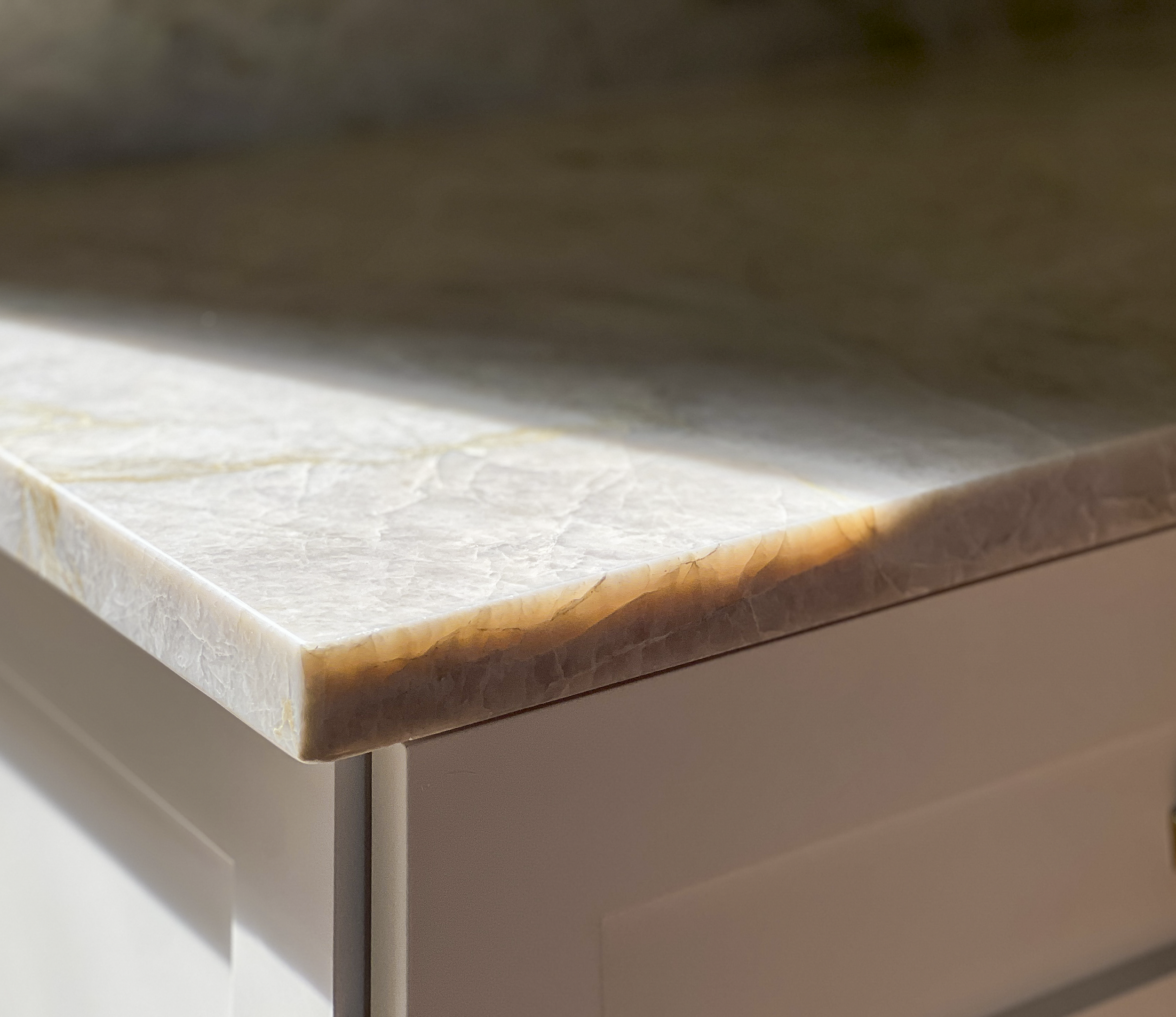 Close-up of a light-colored natural stone countertop edge with subtle gold veining, illuminated by a sharp beam of sunlight on a white shaker cabinet.