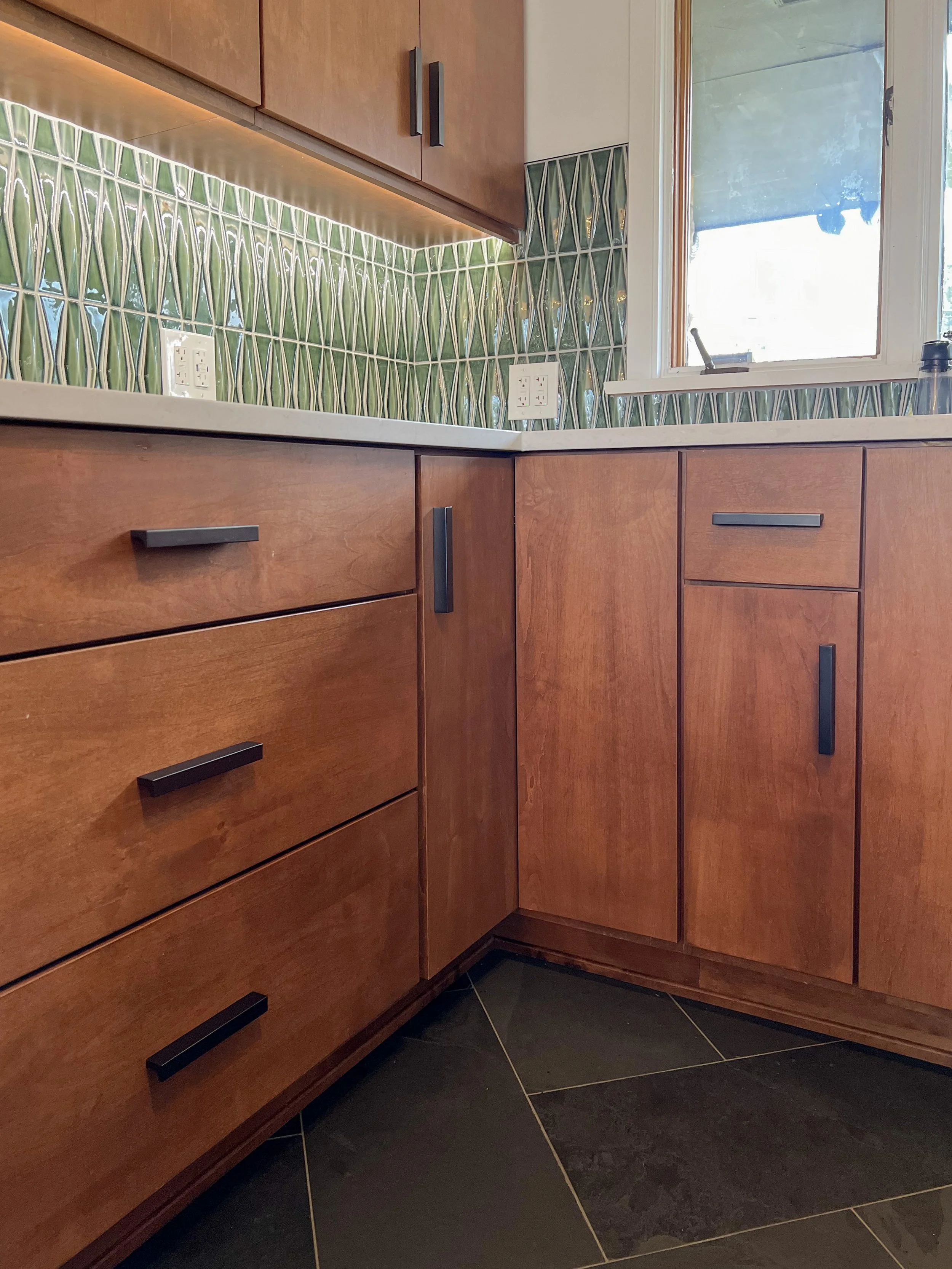 Modern kitchen corner with wooden cabinets and green leaf-patterned tile backsplash, highlighted by under-cabinet lighting.
