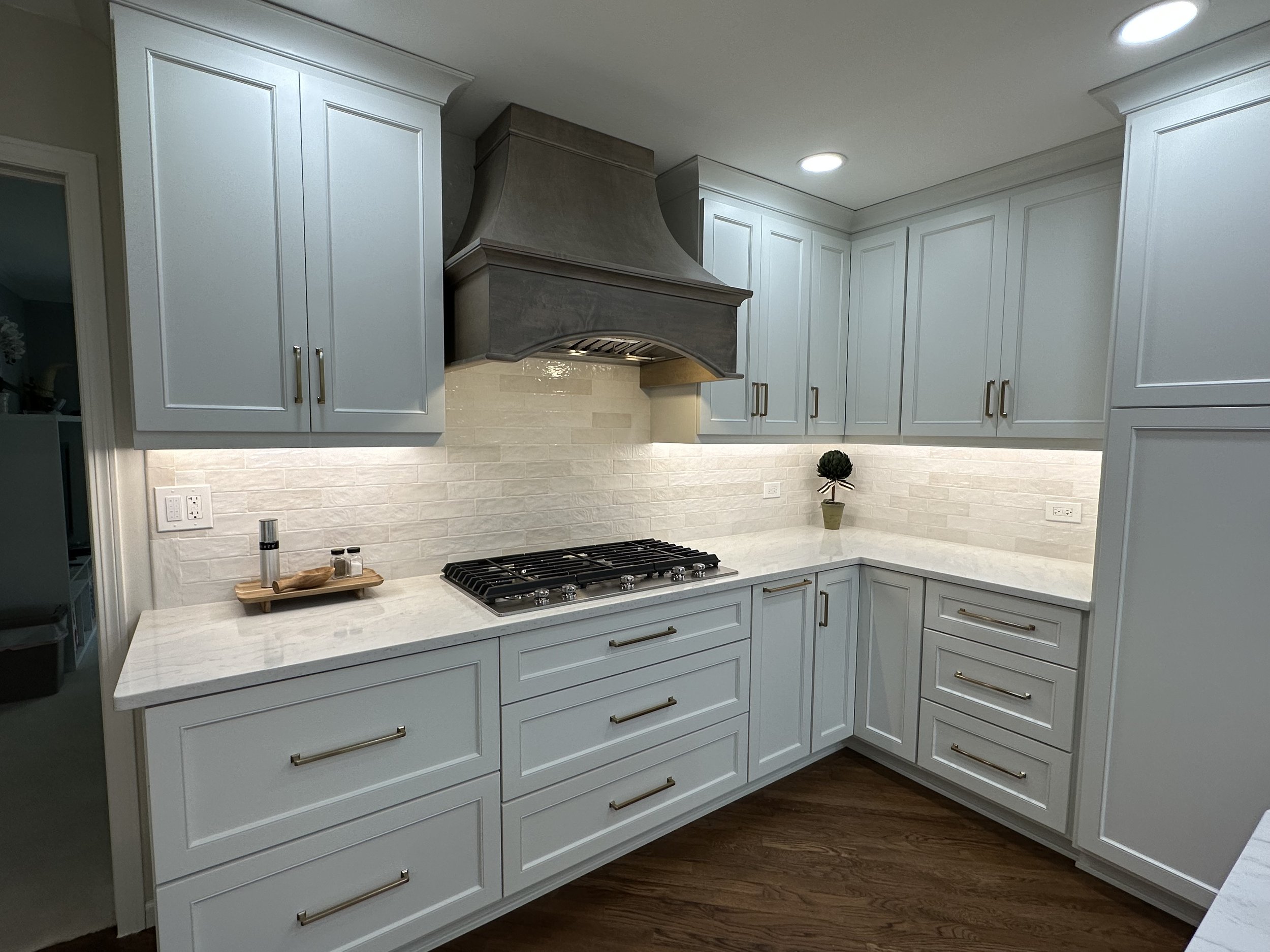 Indulge in the allure of a stunning white kitchen, adorned with a tan stained wood hood, a charming textured subway tile backsplash, and accentuated by honey bronze hardware. A perfect symphony of elegance and warmth, creating a lovely space.