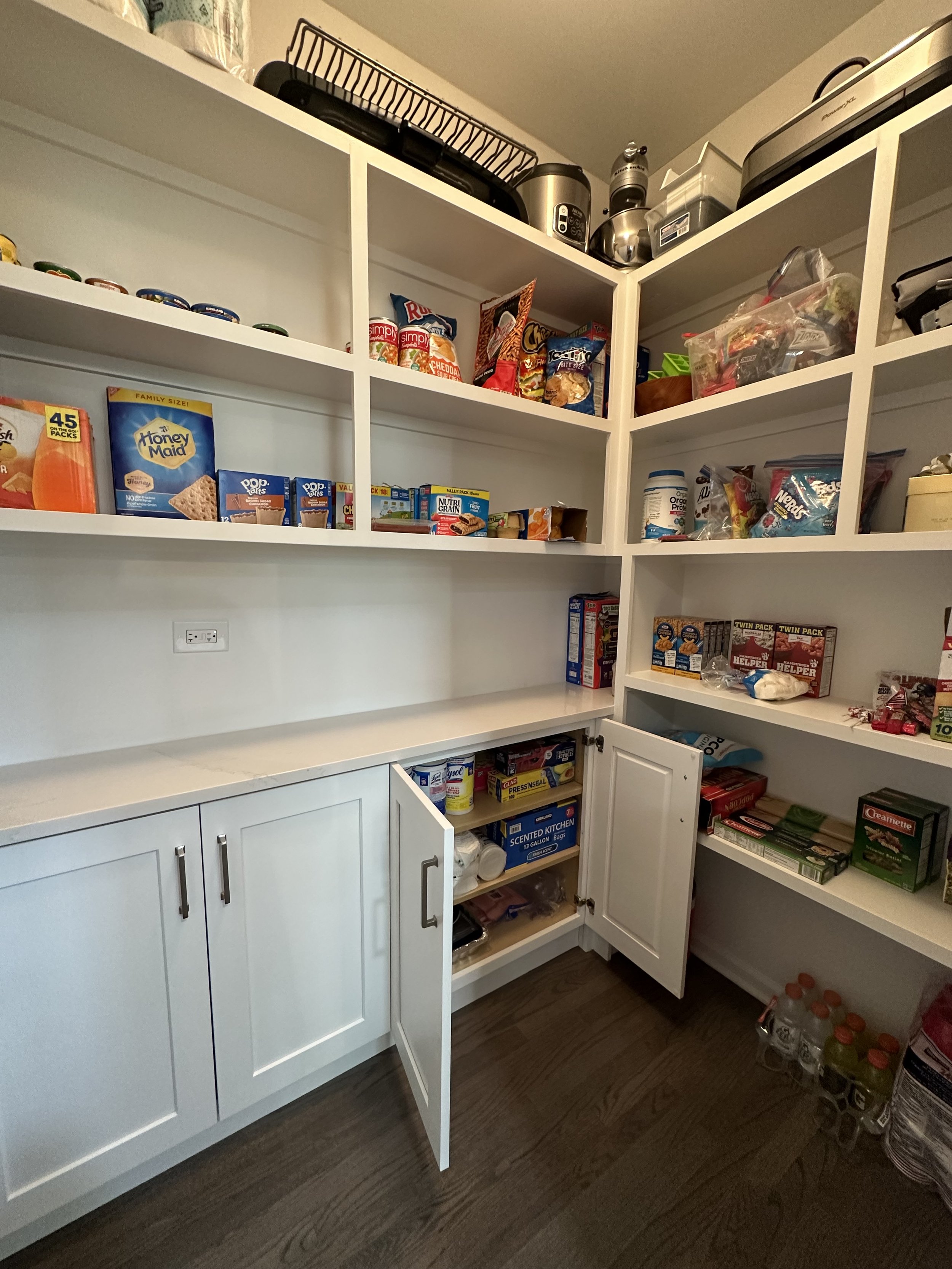 Pantry with shelves containing snacks, canned goods, cereals, appliances, and kitchen supplies. Cabinets below are partially open, revealing more storage items. Bottled beverages are on the floor.
