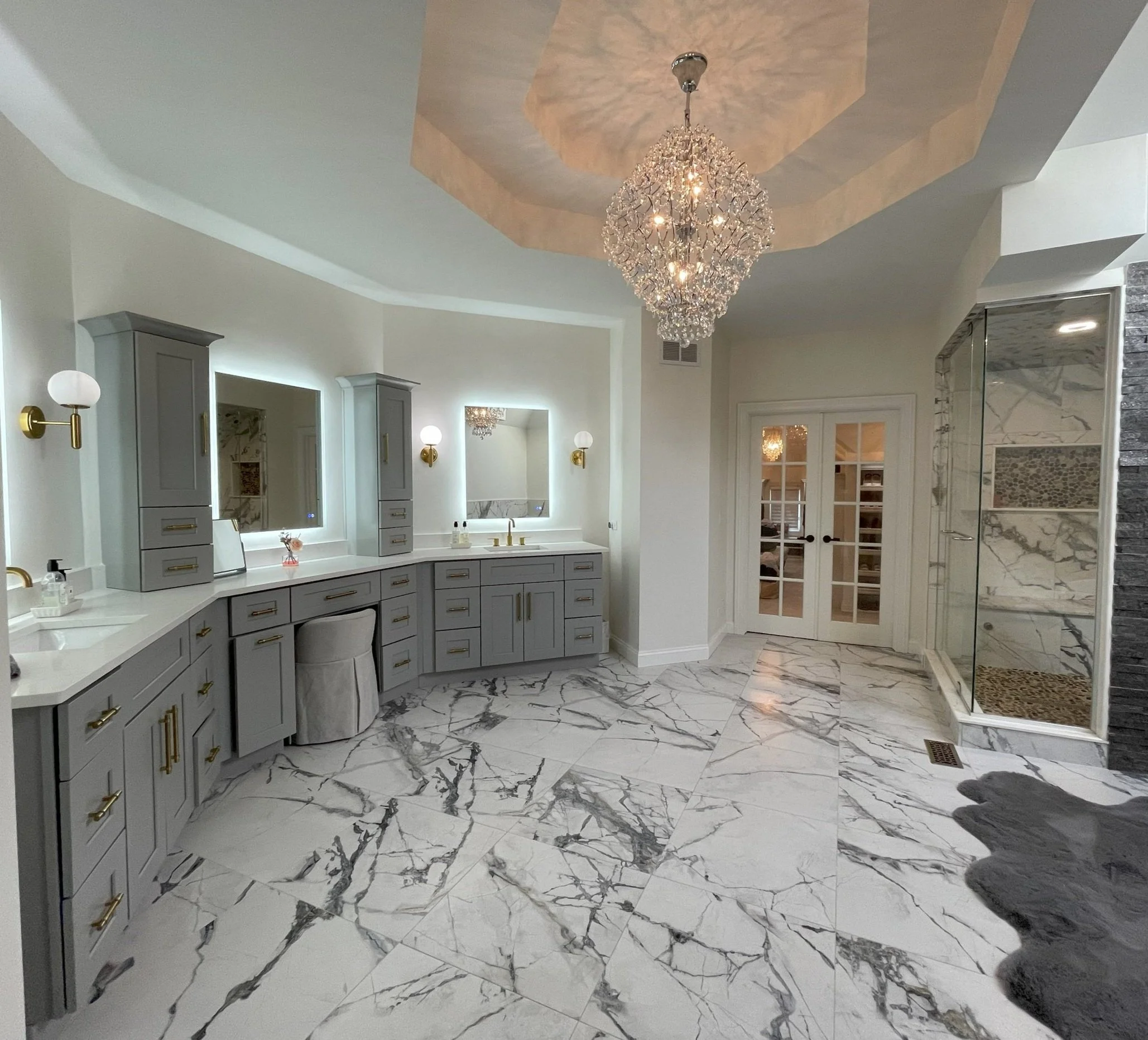 Luxurious bathroom with marble floor, large vanity with dual sinks, mirrored cabinets, crystal chandelier, glass shower enclosure, and double French doors.