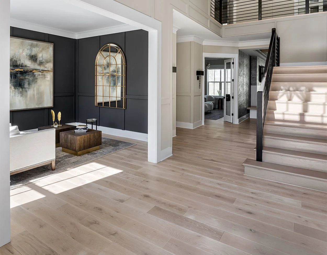 Modern living room with light wood flooring, dark accent wall, abstract painting, arched mirror, wooden coffee table, and a staircase with black railing.