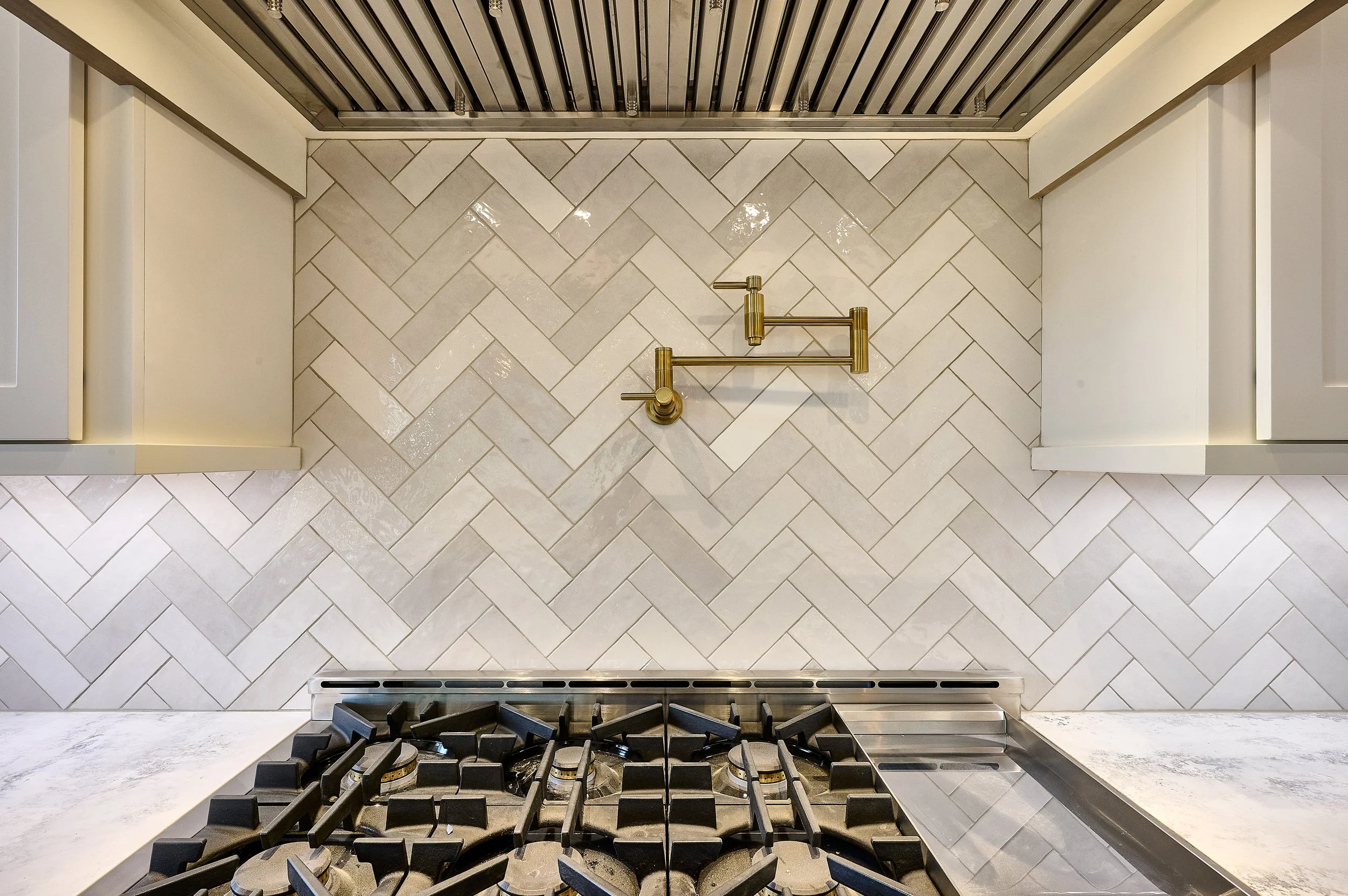 In this captivating photo, a kitchen boasts a chic white and grey tile herringbone backsplash, providing a timeless and visually dynamic backdrop. The luxurious touch of a gold pot filler above the range adds opulence and modern flair to this kitchen
