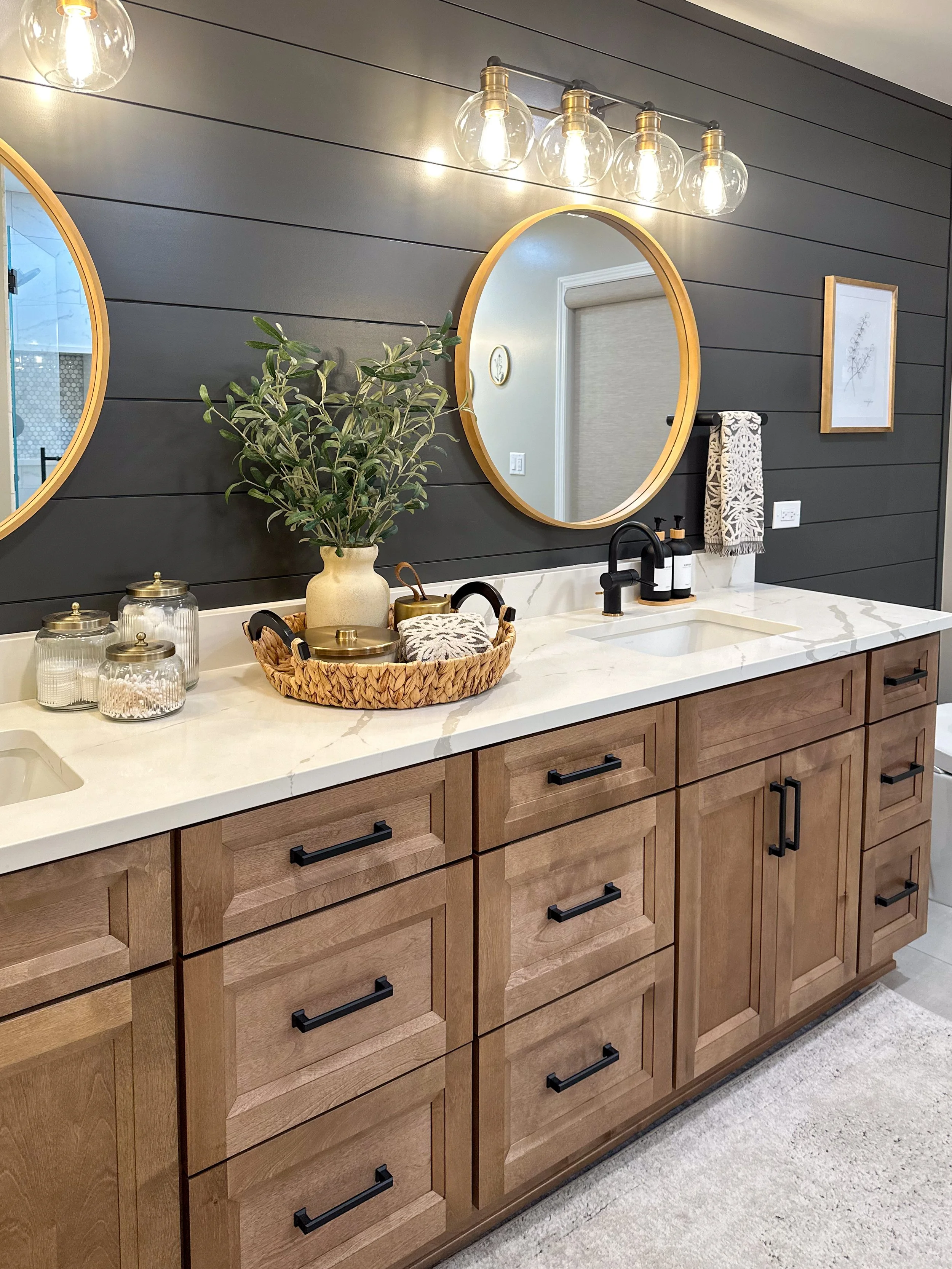 Modern bathroom with wooden vanity and double sinks, round mirrors, and overhead light fixtures. Decor includes a potted plant, a woven basket, and glass jars on the countertop. Dark shiplap wall and tile flooring.