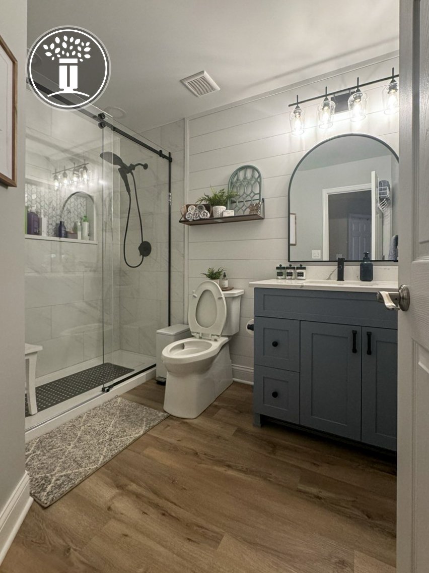 Modern bathroom with a glass shower enclosure, dark fixtures, toilet, blue vanity with double sinks, large mirror, wooden floor, and decorative shelf with plants and towels.