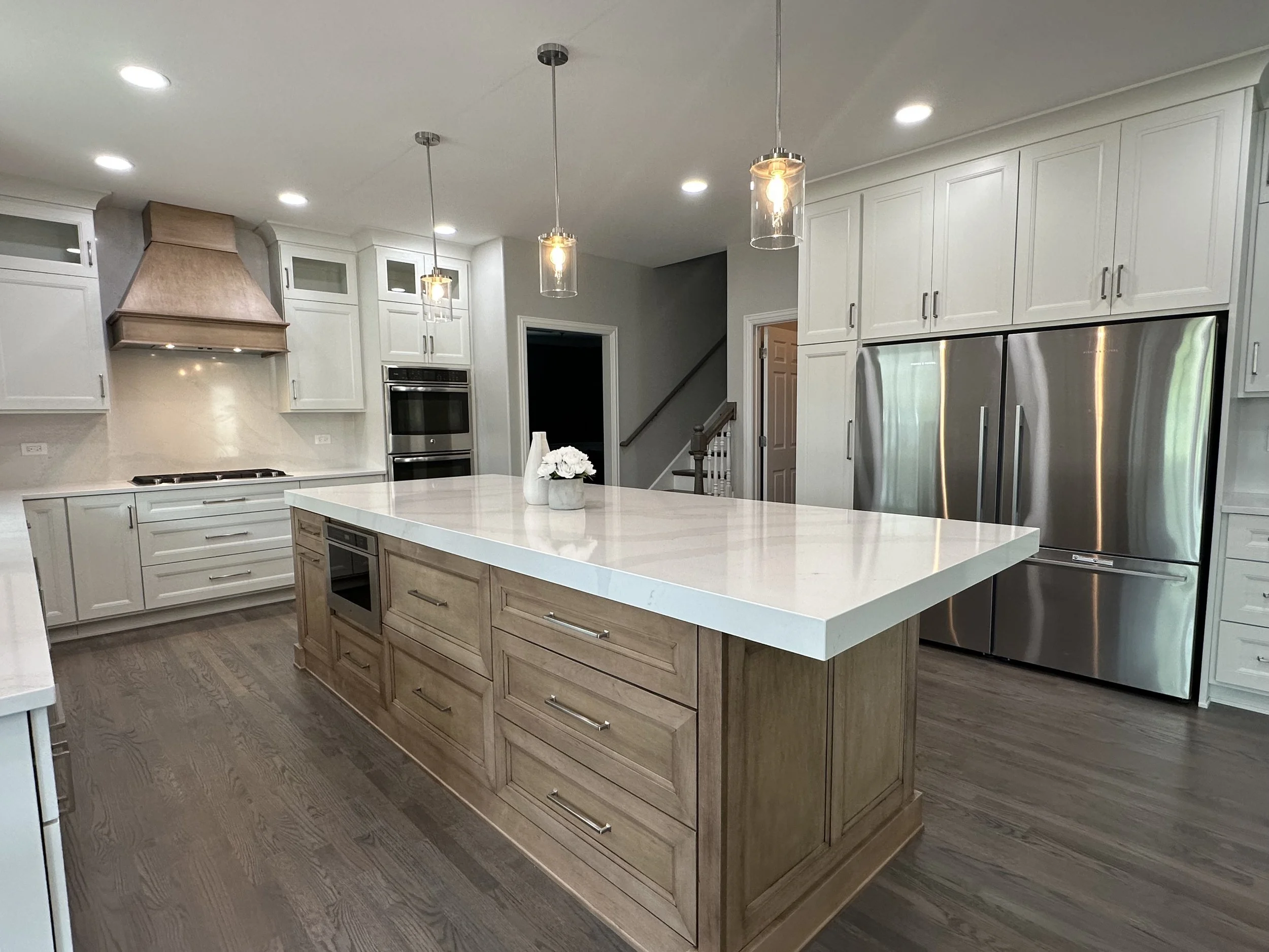 Modern kitchen interior with large white island, stainless steel appliances, pendant lighting, white cabinets, and wooden accents.