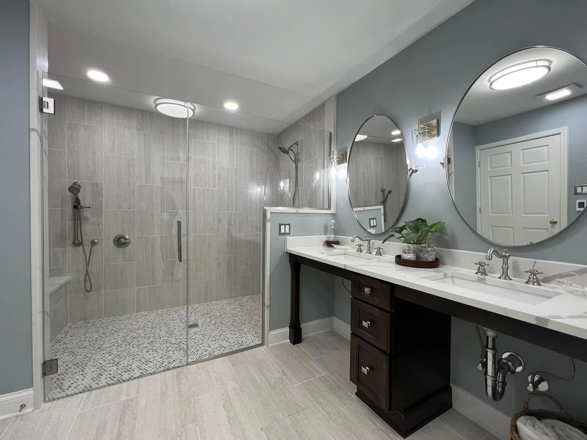 Modern bathroom with a large walk-in glass shower, dual sinks with white counters, and round mirrors. Light gray tiles, dark wood cabinet, and a plant on the countertop.