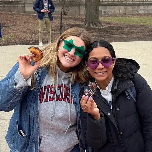 Two women wearing colorful sunglasses smiling and holding donuts on a college campus.