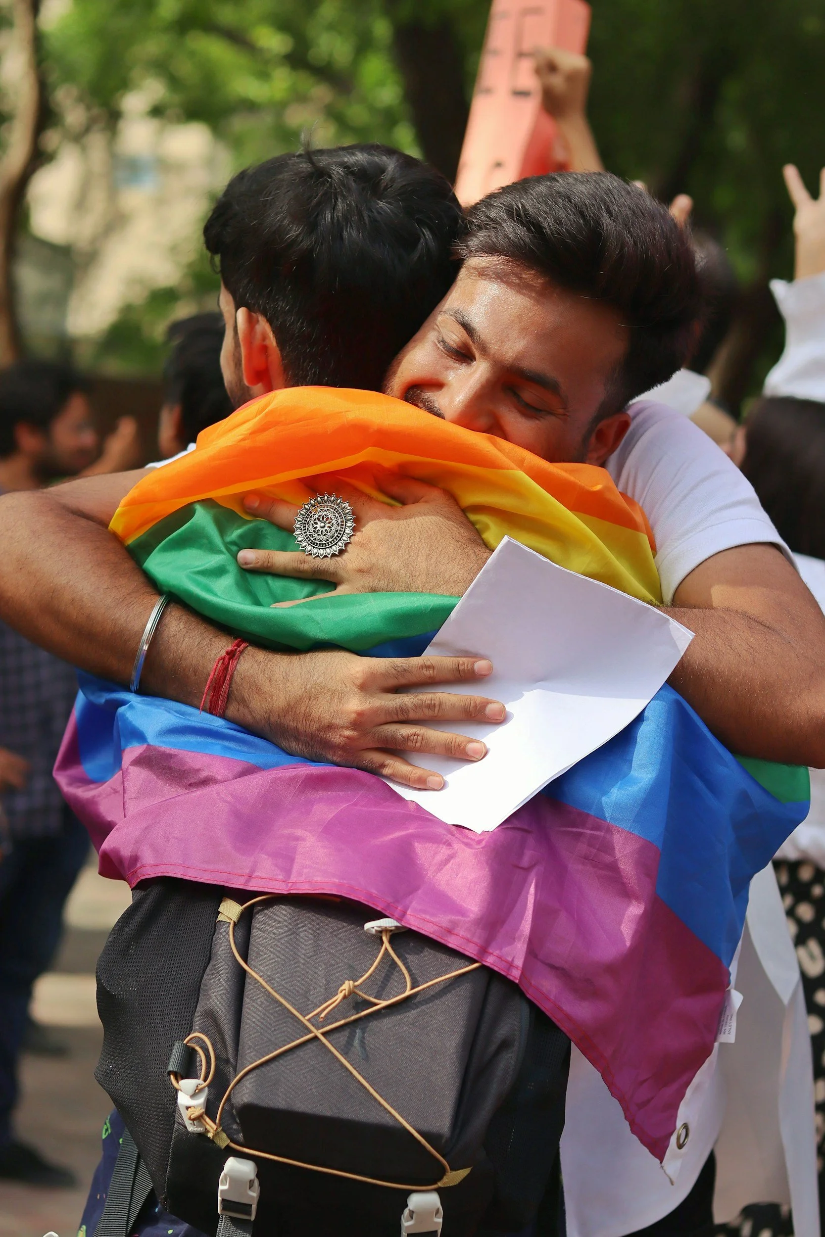 Two men hugging, one wearing a rainbow flag tied around his neck, in a crowd outdoors during daytime.