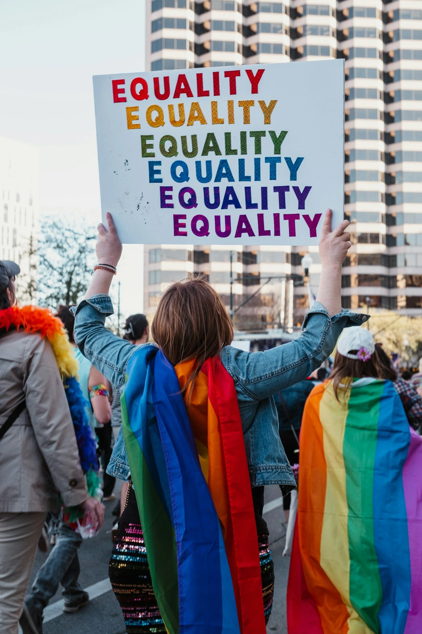 Person holding a sign reading 'EQUALITY' in rainbow colors at a pride parade, with people wearing rainbow flags and colorful clothing in an urban setting.
