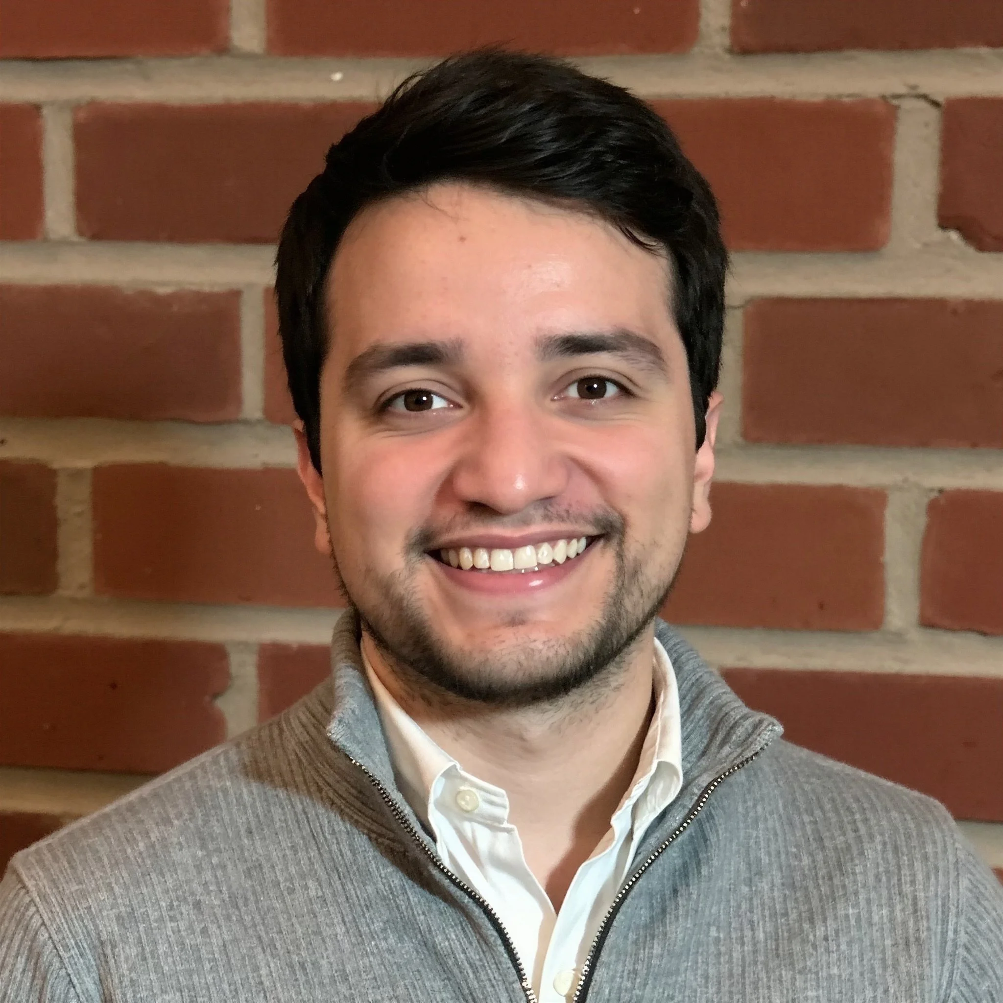 Close-up of a young man smiling, showing his teeth, with short dark hair, wearing a grey zip-up sweater over a white collared shirt, standing in front of a red brick wall.