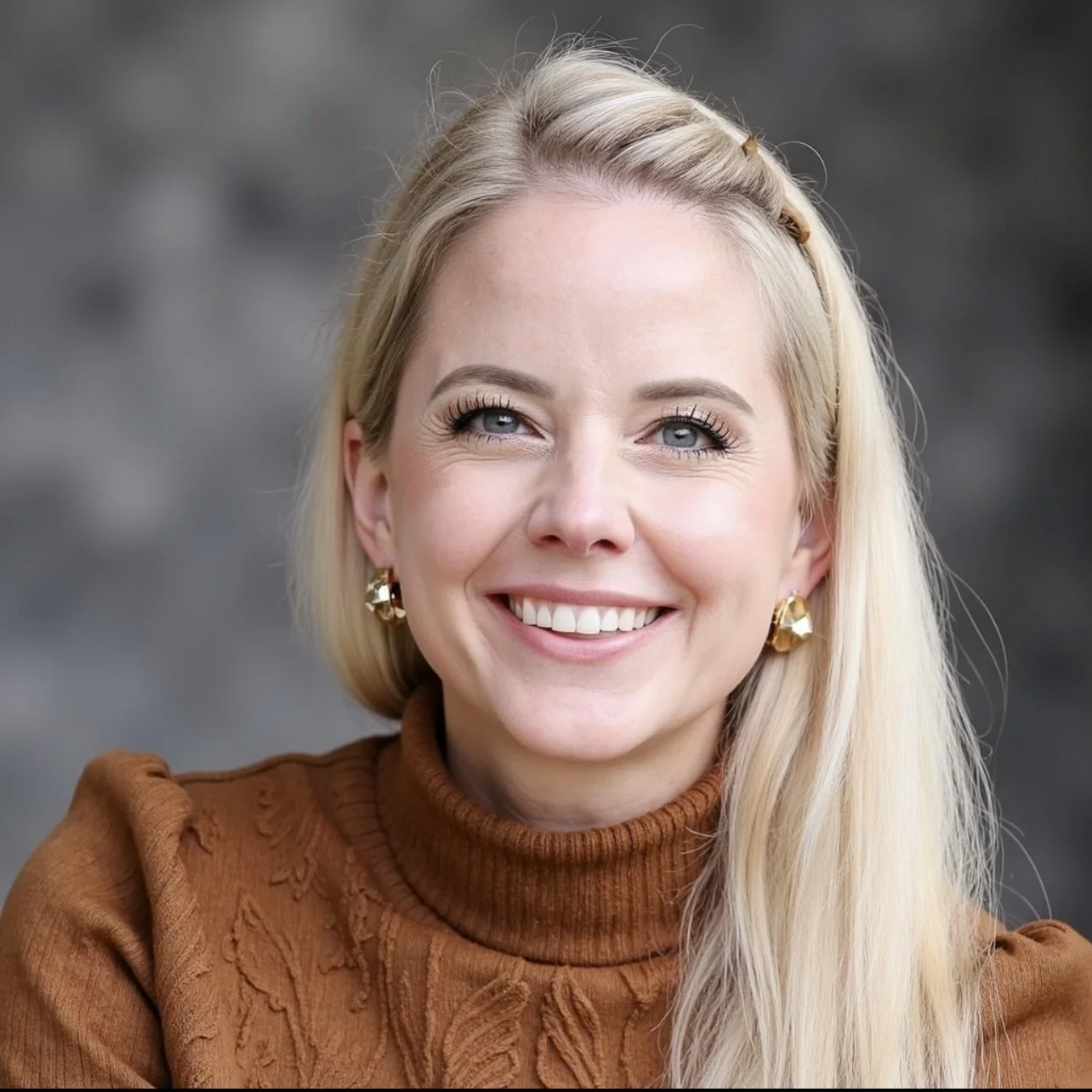 Close-up portrait of a smiling blonde woman with blue eyes, wearing gold earrings and a brown turtleneck sweater, against a blurred gray background.