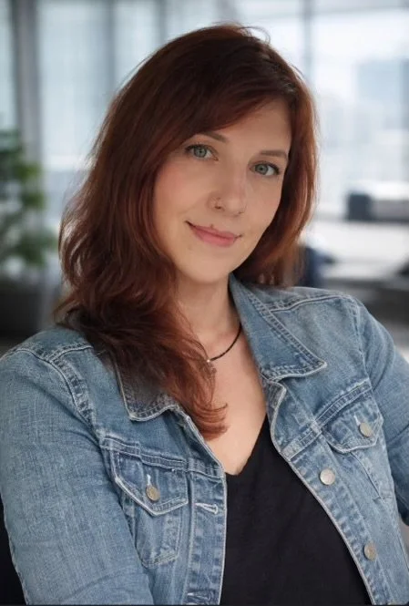 A woman with red hair and blue eyes wearing a denim jacket and black top, smiling in an office setting with large windows.