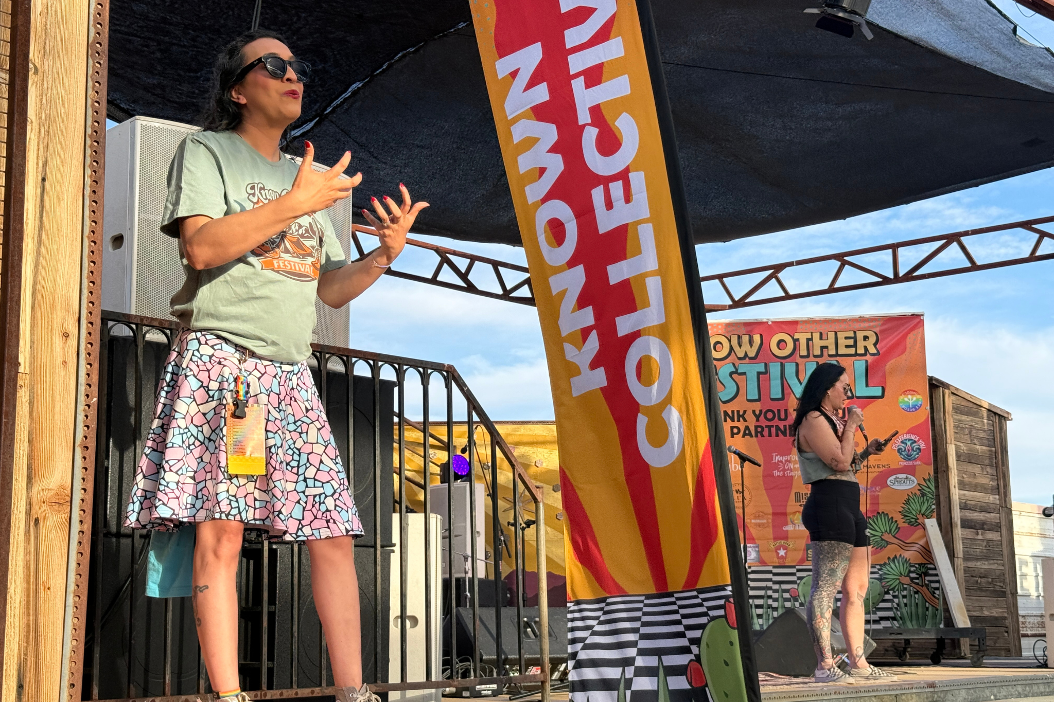 Two women speaking on an outdoor stage at a festival, with one woman in the foreground wearing sunglasses, a graphic T-shirt, and a colorful patterned skirt, and the other woman in the background holding a microphone with a banner and festival signs behind them.
