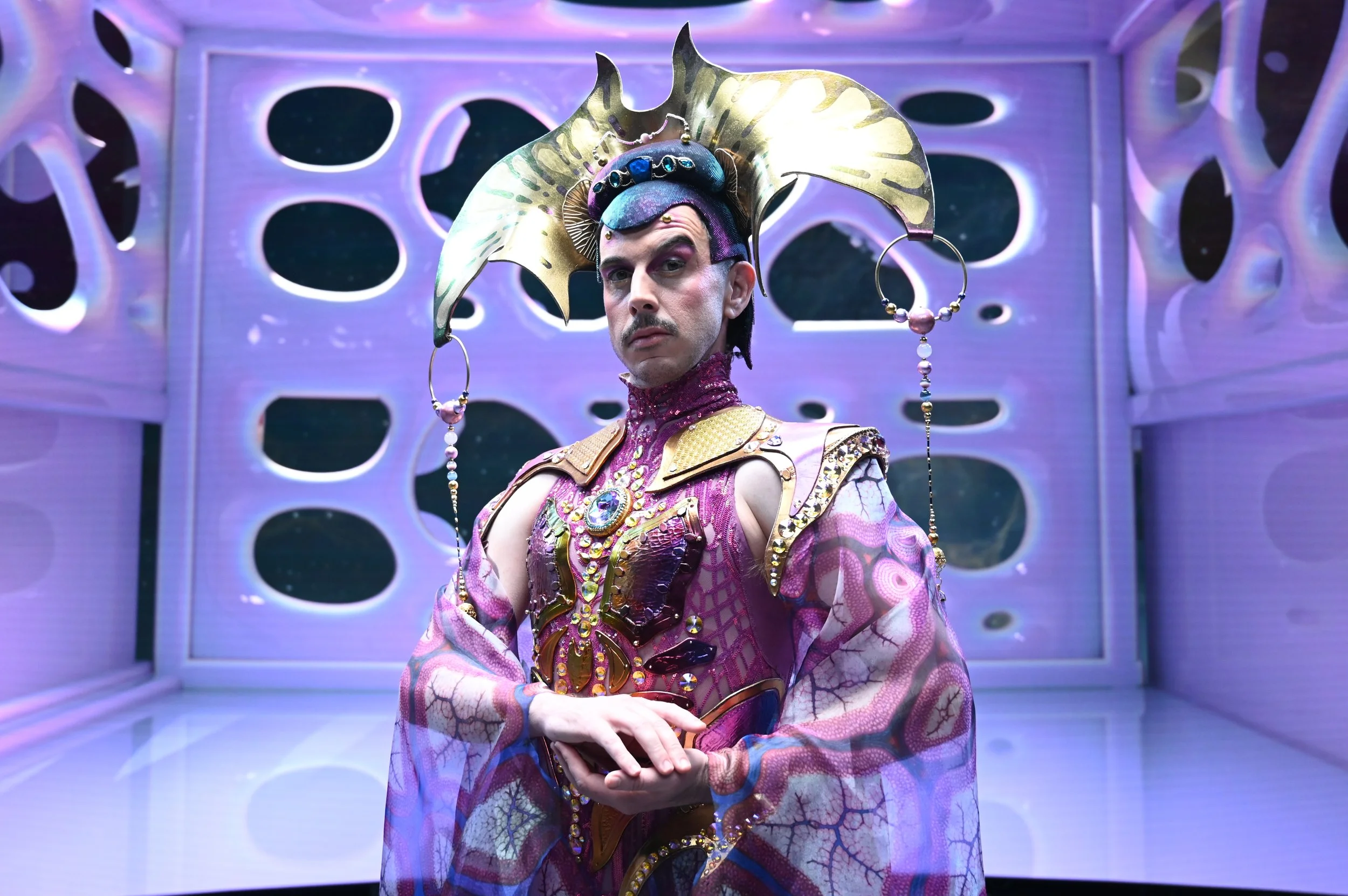 Promotion shot on killjoy. A man wearing a purple dress and golden headpiece stands in a celestial space station.