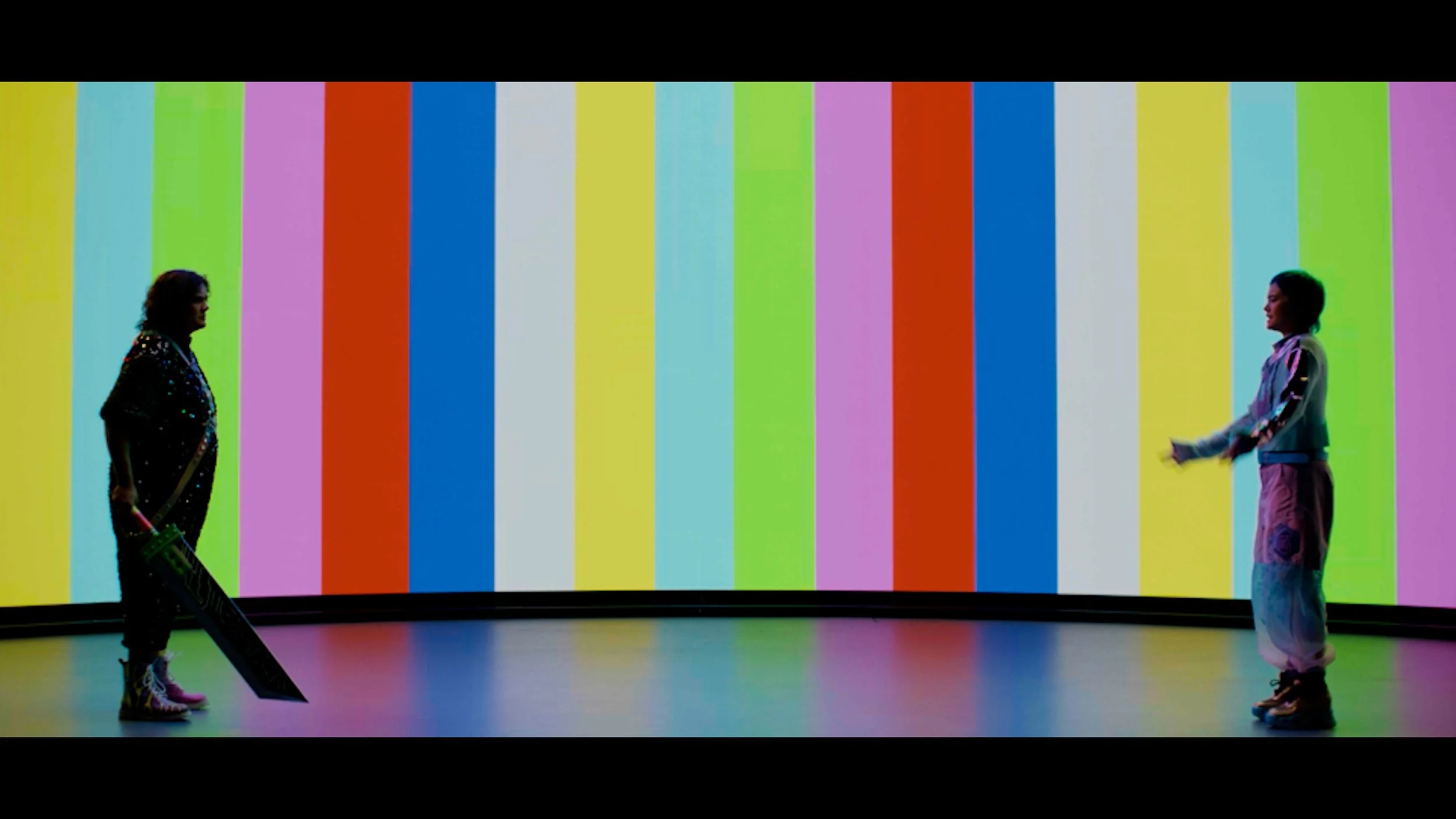 Two non-binary characters facing each other in front of a large, colourful striped screen with vertical rainbow colours.