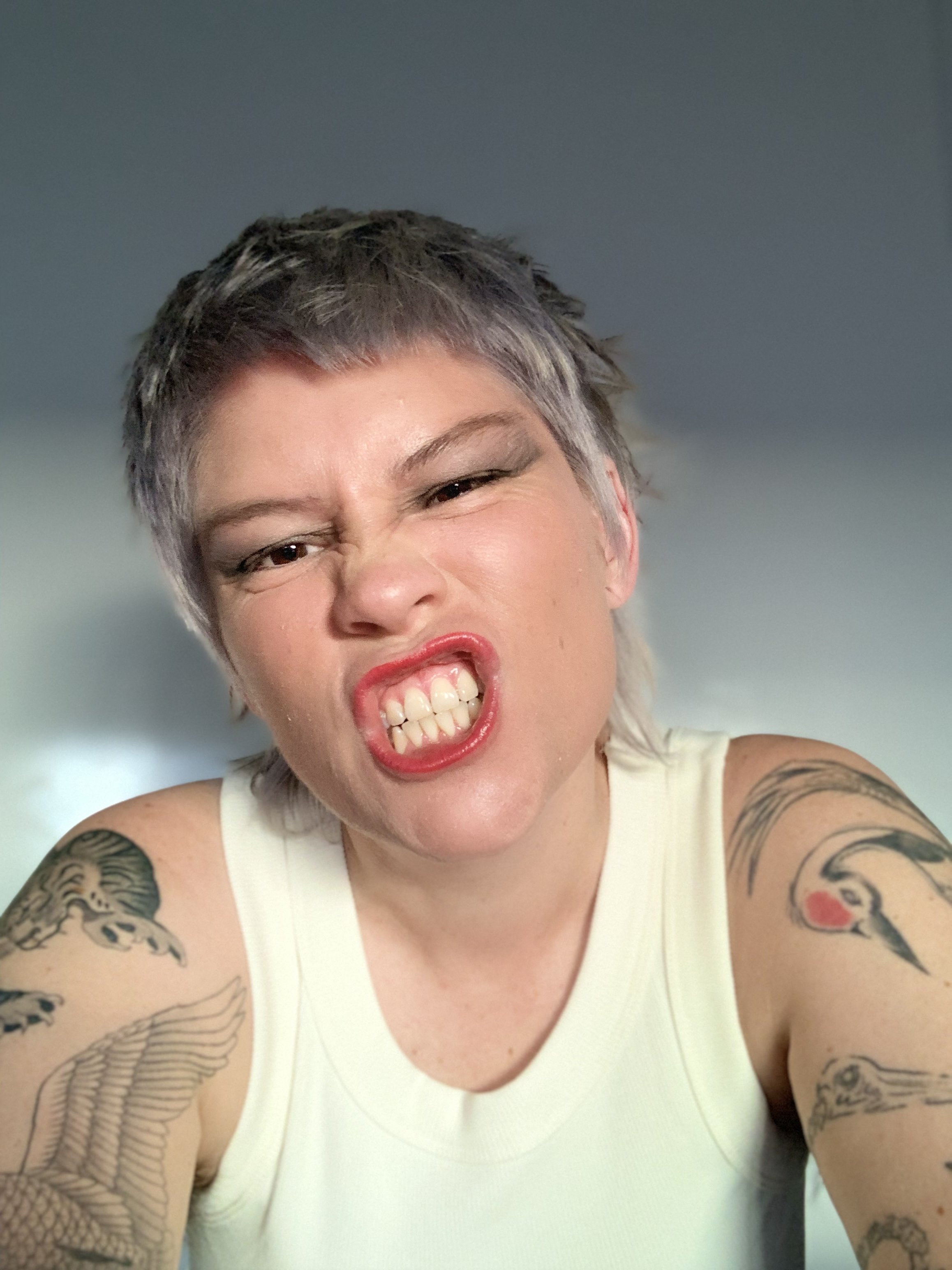 Headshot of director Auryn Ona. They are baring their teeth wearing red lipstick and in white singlet with tattoos on arms.